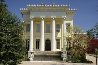 A straight view of Evergreen Museum & Library's front portico. The building is a two-story, yellow Italianate mansion with a two-story, four-columned white portico, topped by ornate cornices. 